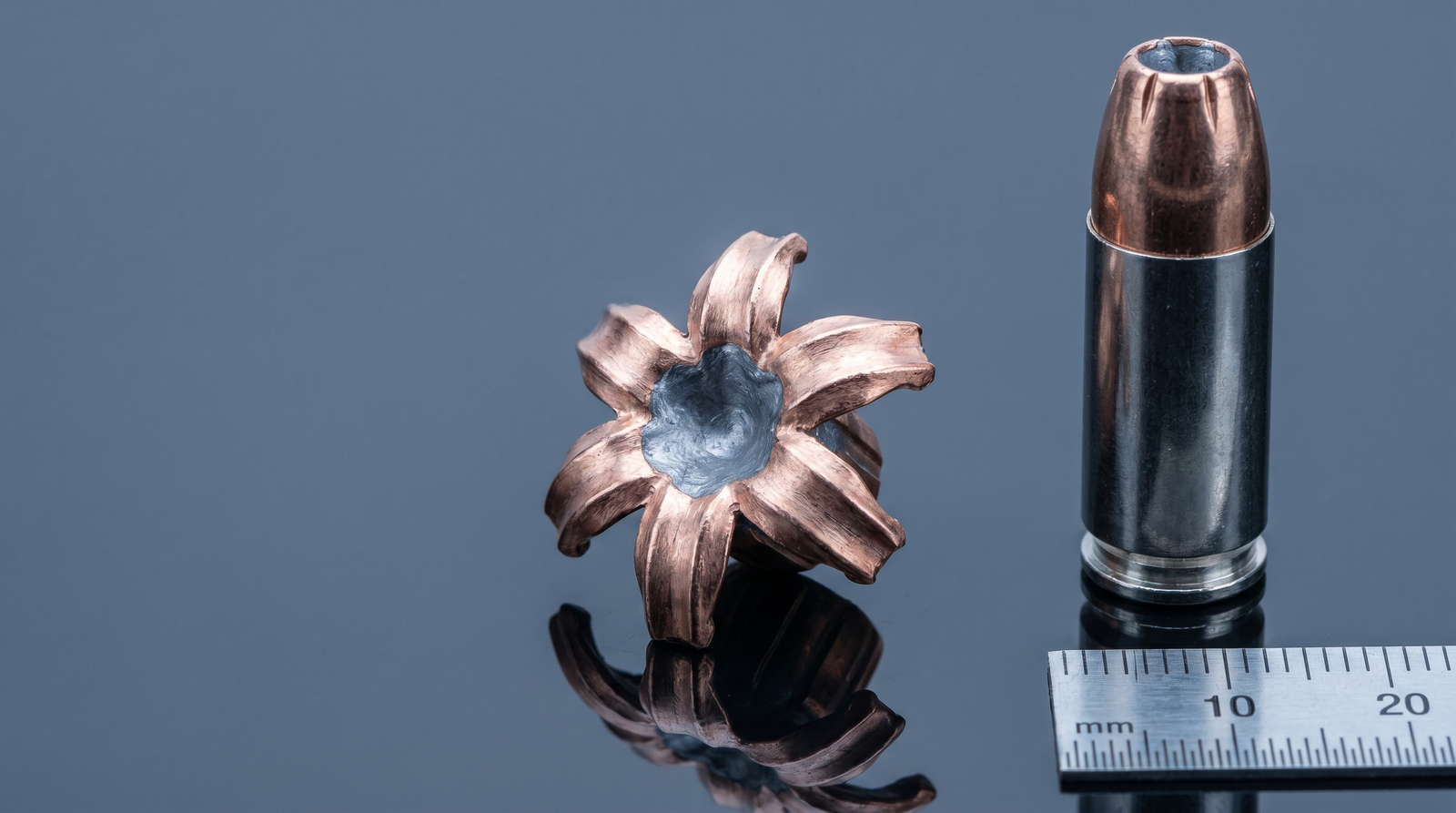 Best 9mm Ammo for Self Defense and Range (2026) 39 Editorial macro photograph of a recovered Federal HST 9mm 124gr jacketed hollow-point bullet expanded into a beautifully mushroomed shape with 6 perfectly-formed copper petals fully splayed open from .355 to roughly .60 caliber expansion alongside an unfired cartridge for comparison and a 10mm metric ruler at frame edge for scale on dark glass studio surface — the FBI-protocol gold standard expansion seen across Federal HST and Speer Gold Dot loads
