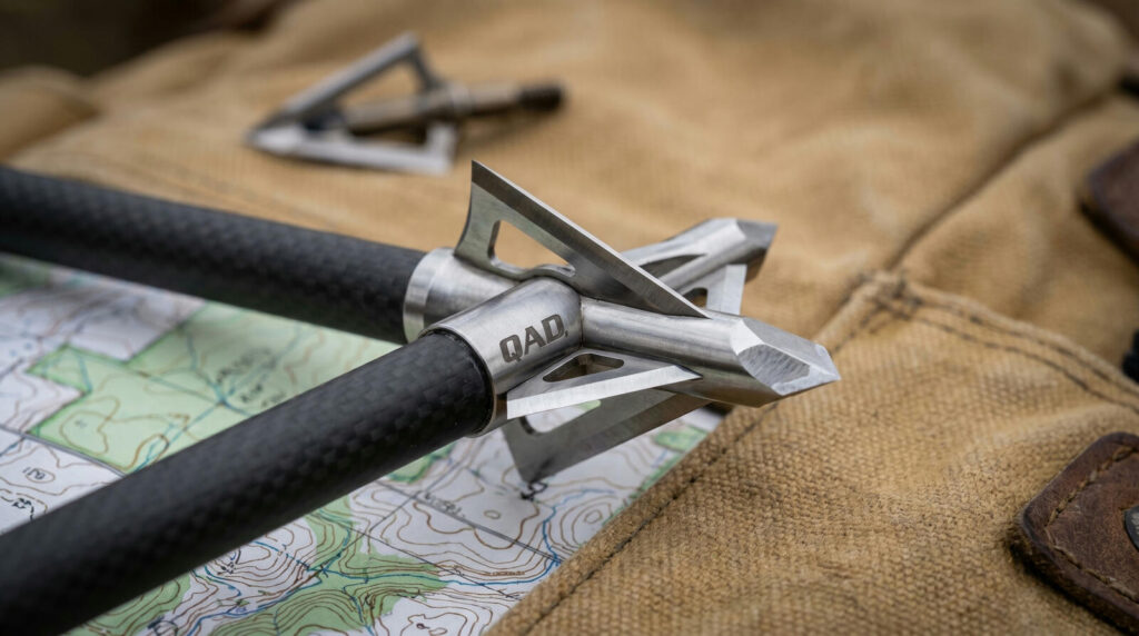 Best Broadheads for Hunting (2026): Fixed-Blade vs Mechanical, Tested 9 QAD Exodus Full Blade four-blade fixed broadhead on carbon arrow on topographic map and tan canvas hunting backpack