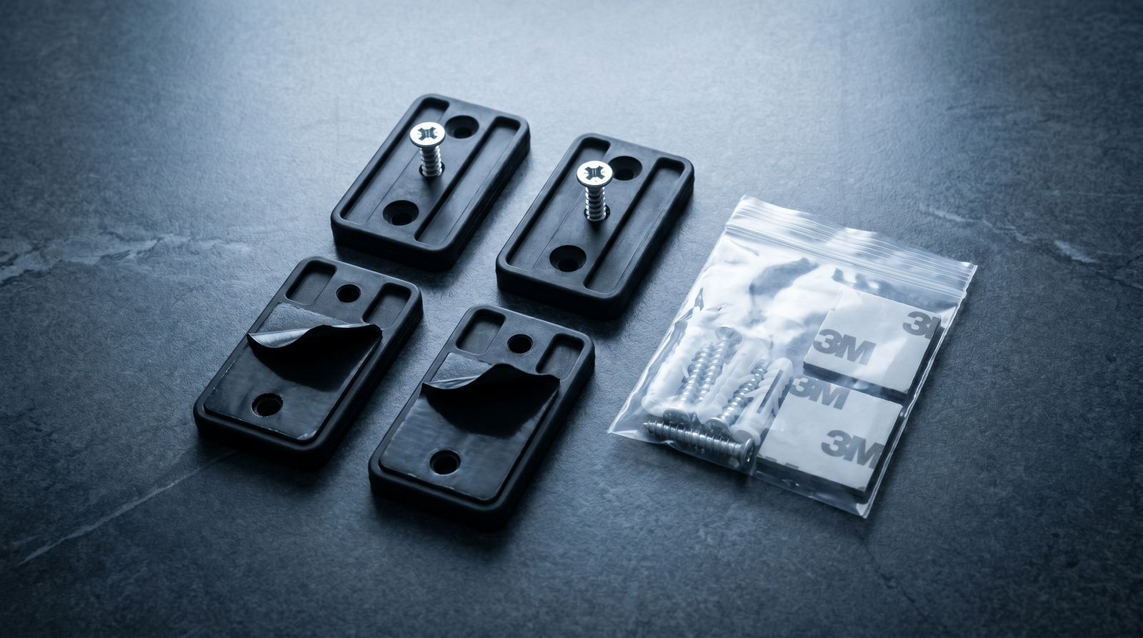 Best Gun Magnets and Quick-Access Mounts (2026) 18 Top-down macro product photograph of four black rubber-coated neodymium gun magnet mounts in a RYMMES-style multi-pack on polished dark slate with two units showing inserted Phillips mounting screws, two units showing peeled 3M VHB adhesive strips, plus a bag of additional mounting screws and 3M adhesive squares under cool overhead LED studio lighting