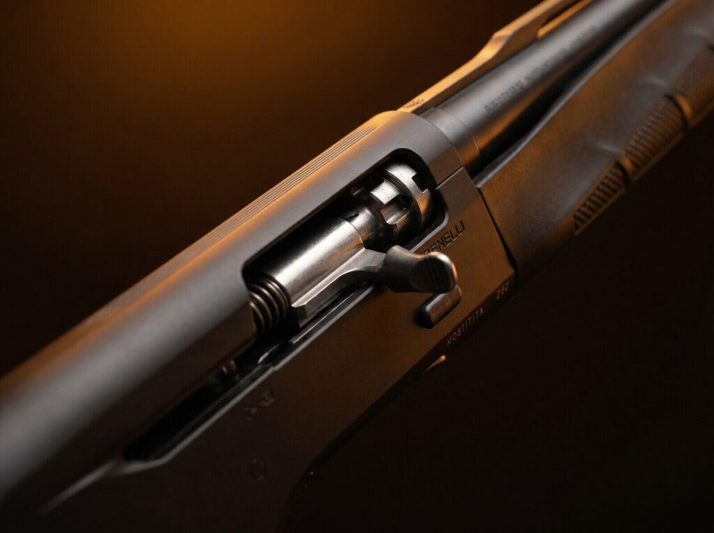 Benelli M2 Review (2026): 500 Round Test of the Inertia-Driven Legend 13 Macro close-up of the inertia-driven bolt system on the Benelli M2 Field 12 gauge showing the silver bolt face, BENELLI receiver engraving, and recoil spring tube in warm tungsten side-rim lighting