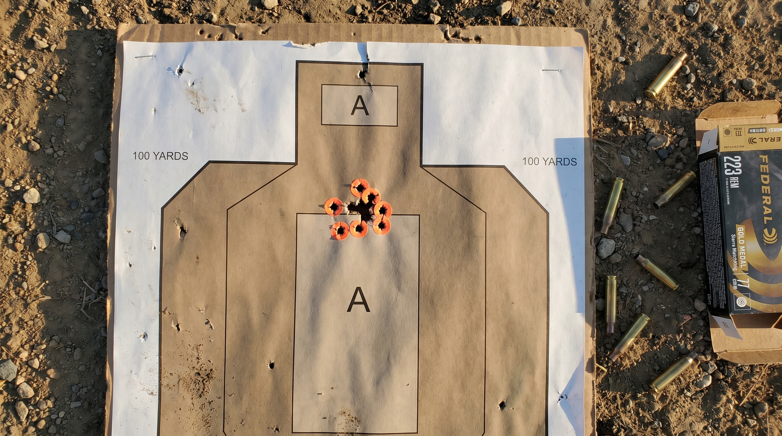 Sig MCX Spear LT Review (2026): 500 Round Test of the Next-Gen AR Alternative 6 Top-down photograph of paper IPSC silhouette target at 100 yards with tight 1.5 MOA five-shot group above the A-zone marked with neon orange shot stickers, scattered spent 5.56 brass and Federal Premium 223 Gold Medal Match ammunition box on outdoor dirt range ground in late-afternoon side-light