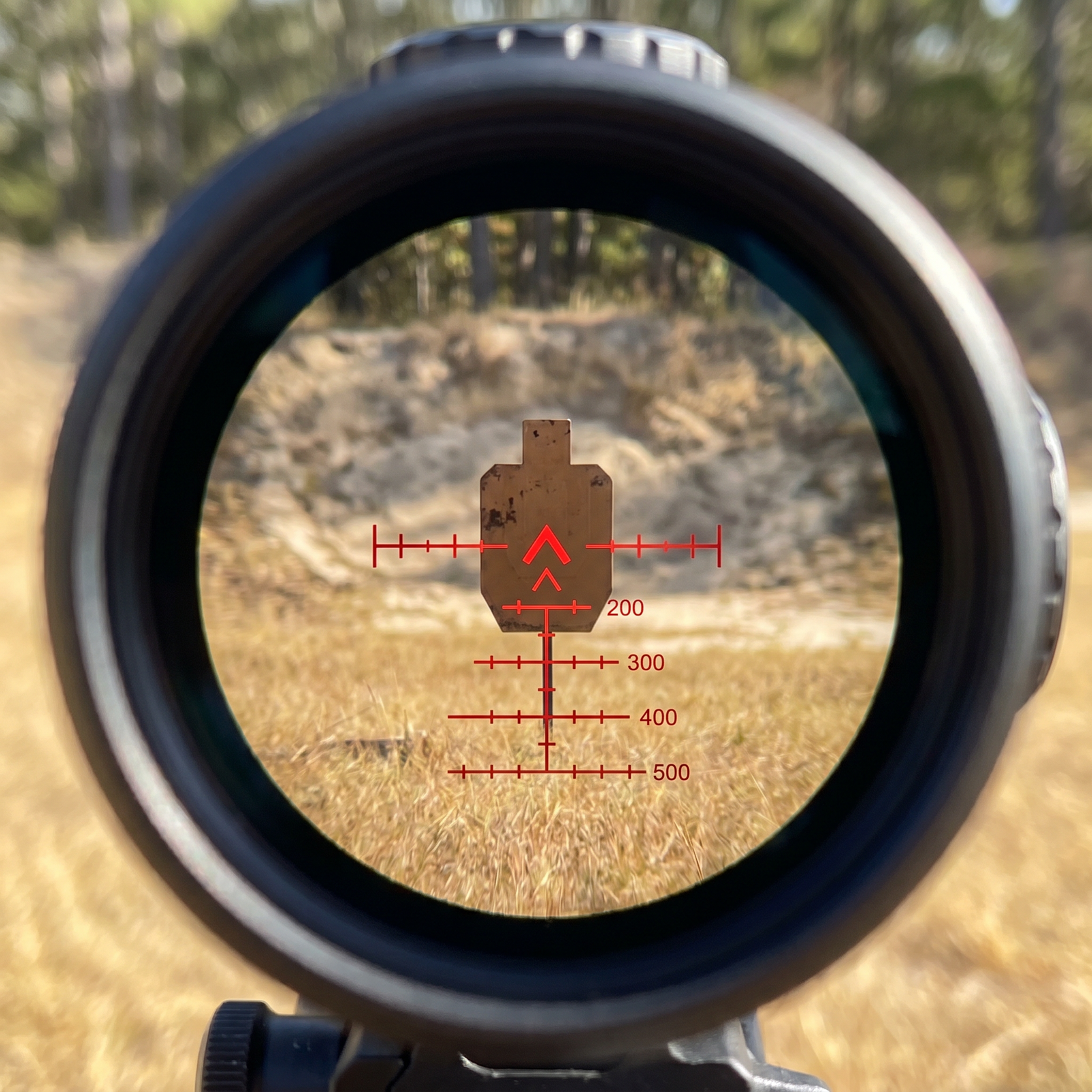 8 Best Prism Scopes in 2026: Tested and Ranked 30 First-person through-the-scope POV view of the Primary Arms SLx 3x MicroPrism ACSS Raptor BDC reticle illuminated in red showing center chevron stadiometric ranging marks and 200/300/400/500-yard BDC hash marks overlaid on outdoor rifle range scene with brown silhouette steel target at 100 yards, dry golden grass foreground and sand berm tree-line backdrop in bright daylight