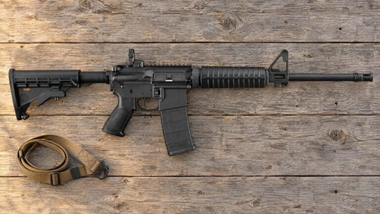 Ruger AR-556 rifle with folding rear sight and two-piece handguard lying flat on weathered barn wood beside a tan sling