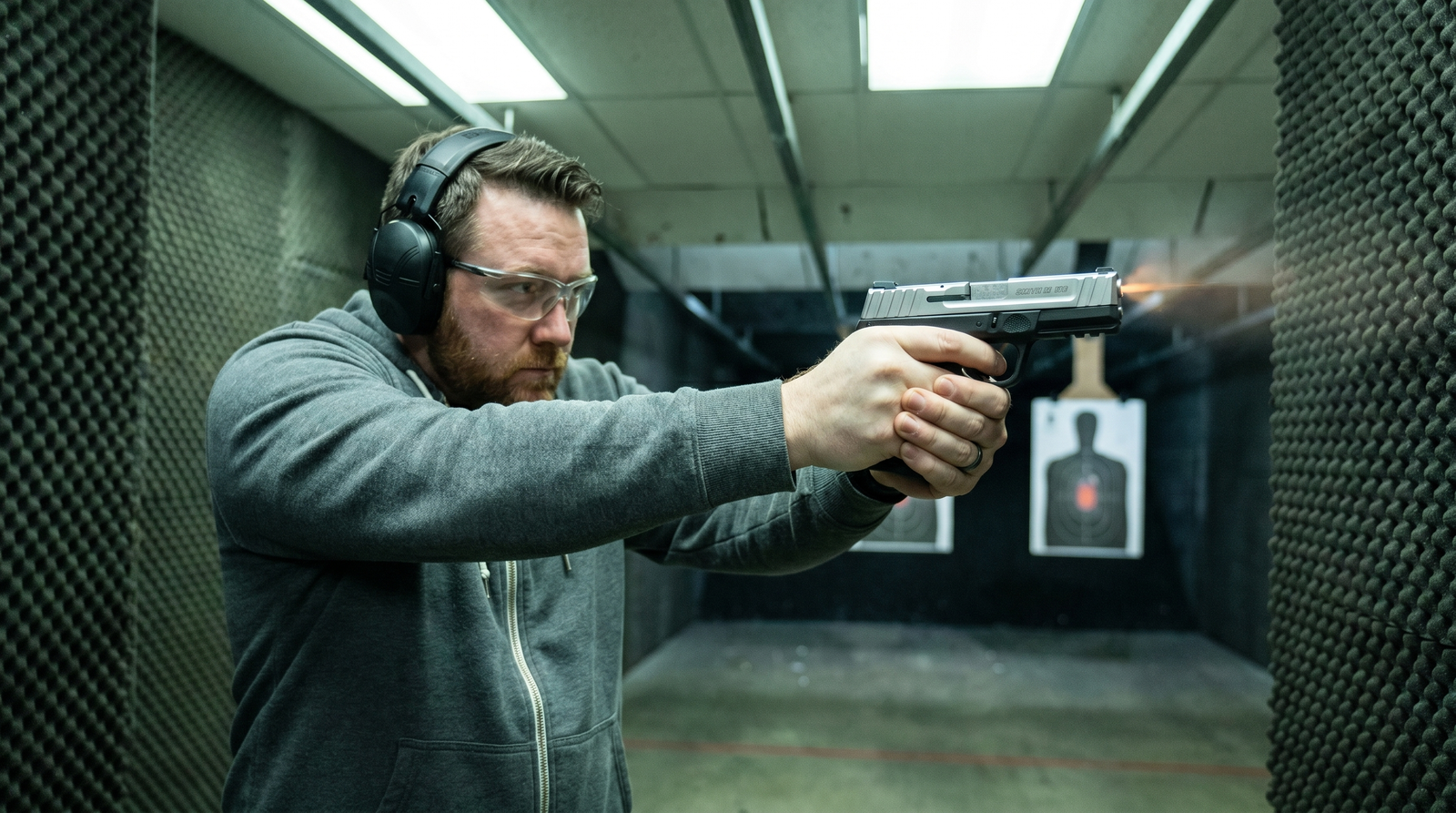 S&W SD9 VE Review (2026): 500 Round Test of the Cheapest Smith on the Shelf 5 Male shooter in grey hoodie and ear pro shooting Smith and Wesson SD9 VE in indoor pistol bay with foam acoustic walls and silhouette targets at 7 yards