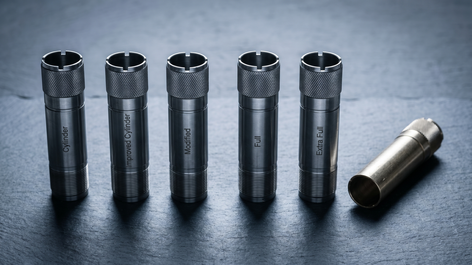 Best Shotgun Upgrades in 2026: Accessories That Actually Matter 39 Studio macro product photograph of five Carlson stainless steel shotgun choke tubes (Cylinder, Improved Cylinder, Modified, Full, Extra Full) standing vertically in a precise row on cool dark grey slate surface with brass choke wrench at right under cool overhead LED studio lighting with hard shadows