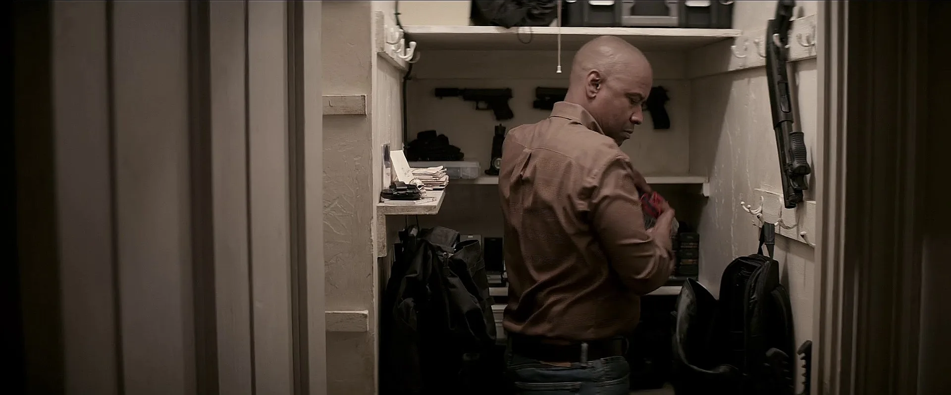 Robert McCall standing in his hidden closet weapons cache with pistols and a short shotgun on the walls in The Equalizer