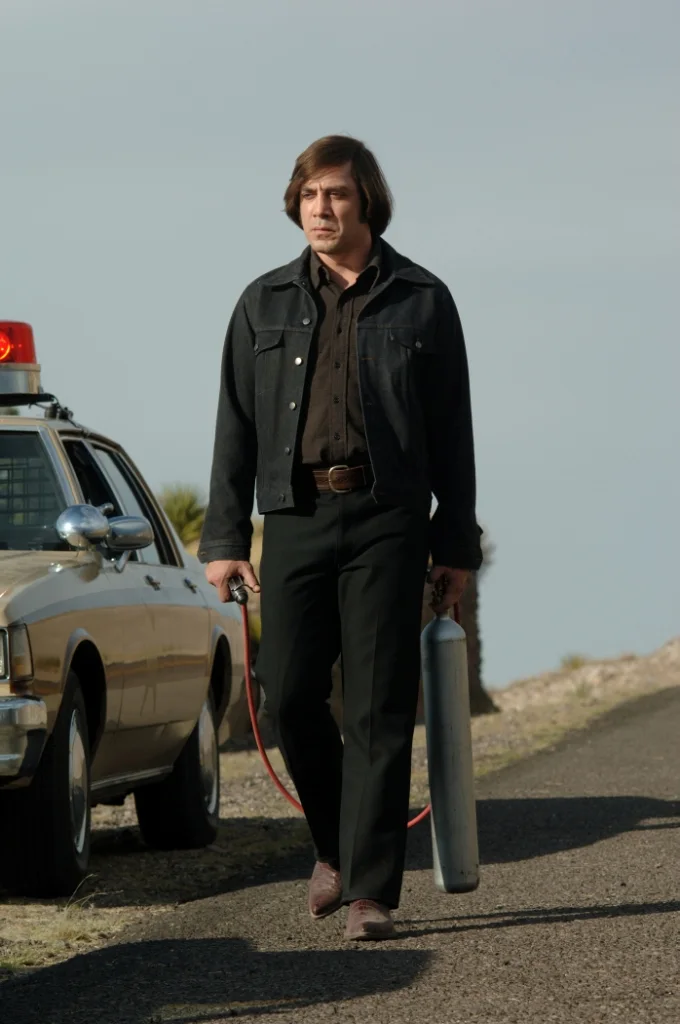 Anton Chigurh walking from a stolen police car carrying a captive bolt pistol and compressed air tank in No Country for Old Men