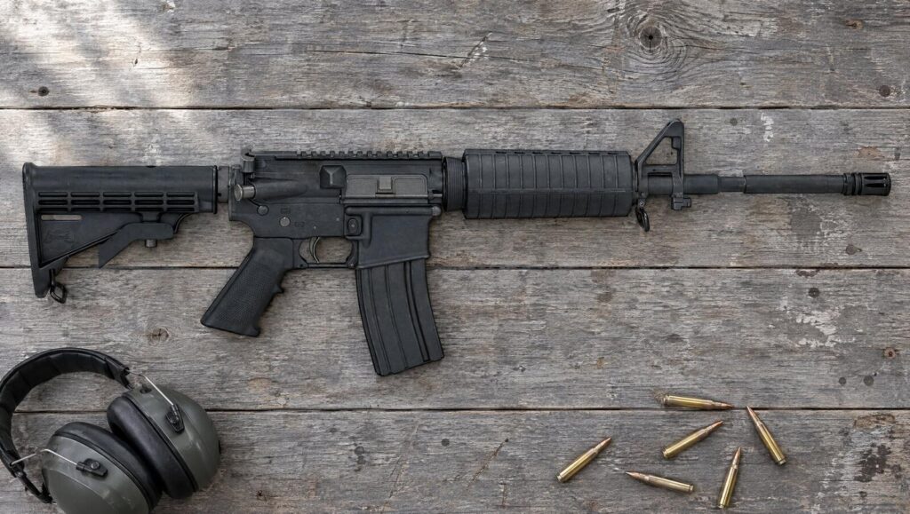A PSA PA-15 M4 carbine AR-15 lying flat on a gray barn-wood workbench beside ear muffs and loose 5.56 rounds