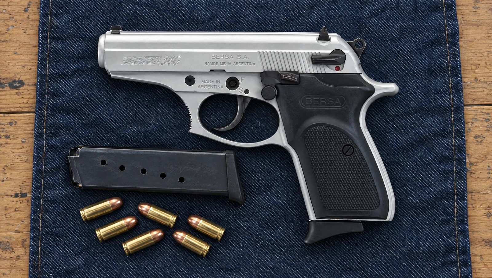 Bersa Thunder 380 pistol in satin nickel with black grips lying on blue denim beside a spare magazine and six .380 ACP cartridges