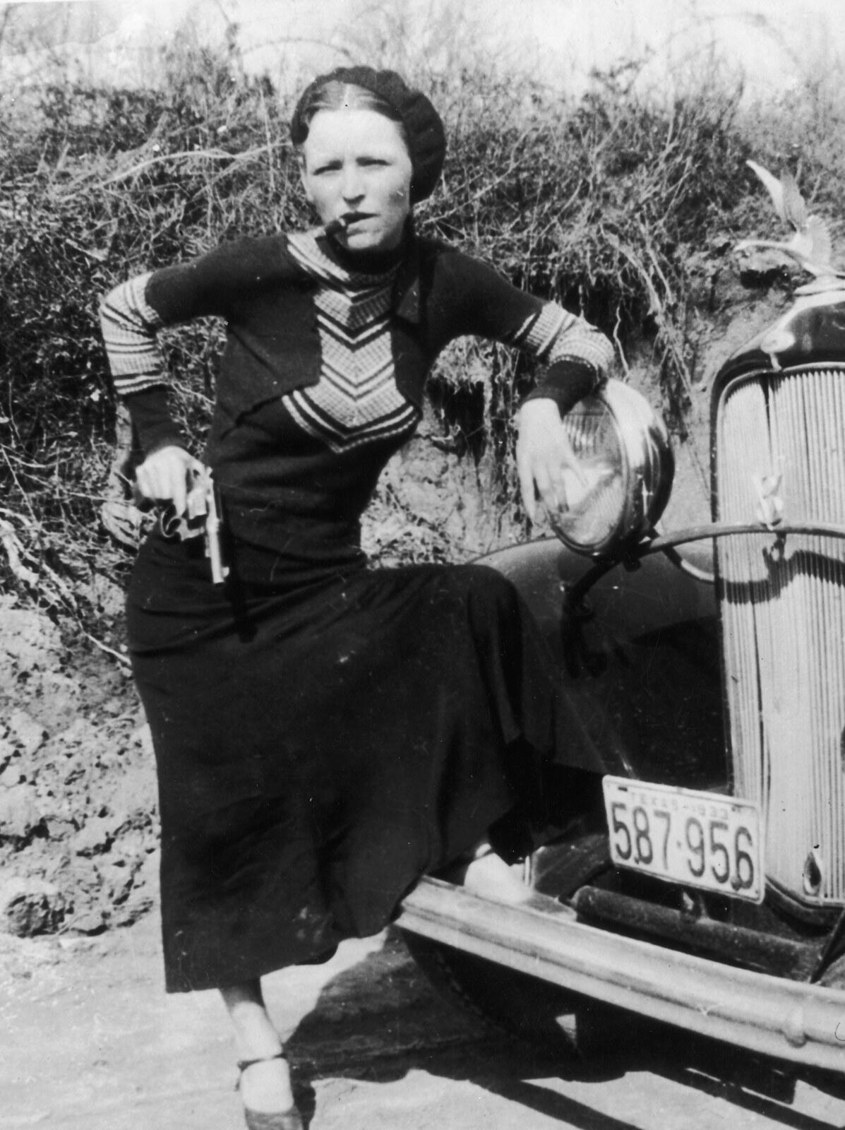 Bonnie Parker posing with a cigar and a revolver against a car in 1933