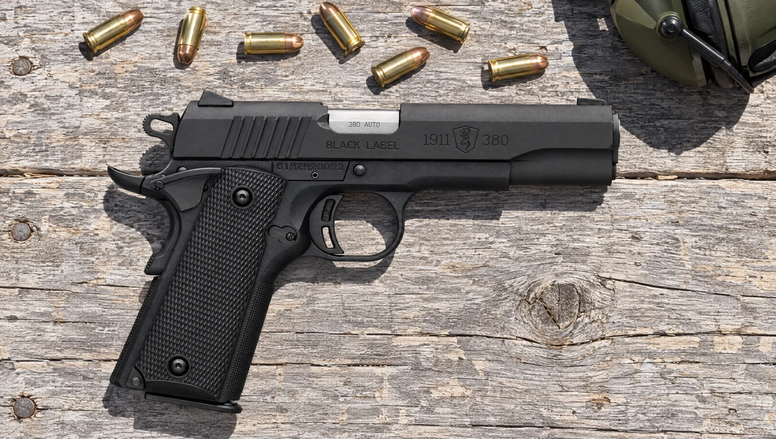 Browning 1911-380 Black Label .380 ACP pistol lying on a weathered wooden range bench in bright sun beside loose .380 ACP cartridges and hearing protection