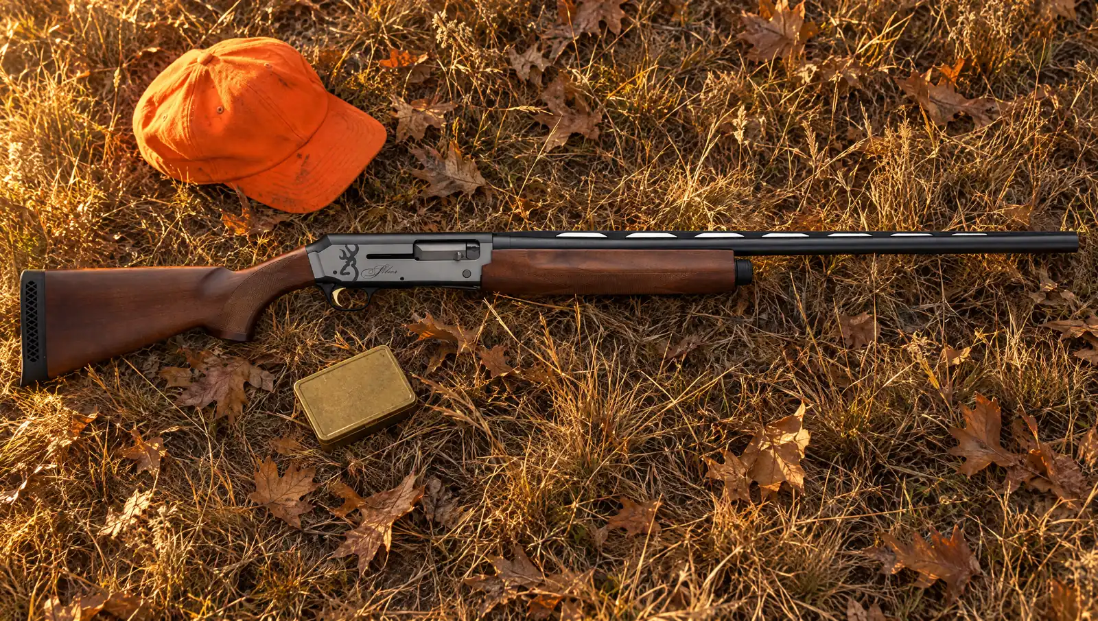Browning Silver Field 12 gauge with a satin Turkish walnut stock and a gray and black bi-tone receiver lying in dry autumn grass and oak leaves beside a blaze orange hunting cap and a brass shell box