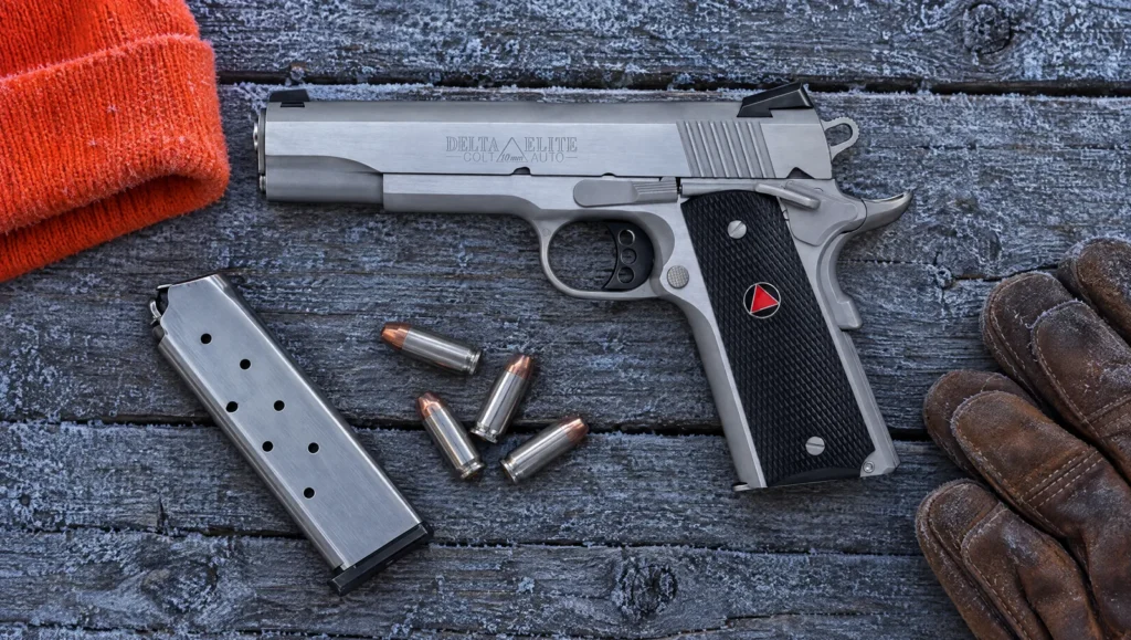 Left side of a stainless Colt Delta Elite 10mm marked Delta Elite Colt 10mm Auto, lying on frosty boards with a spare magazine, 10mm cartridges, an orange hunting cap and leather gloves