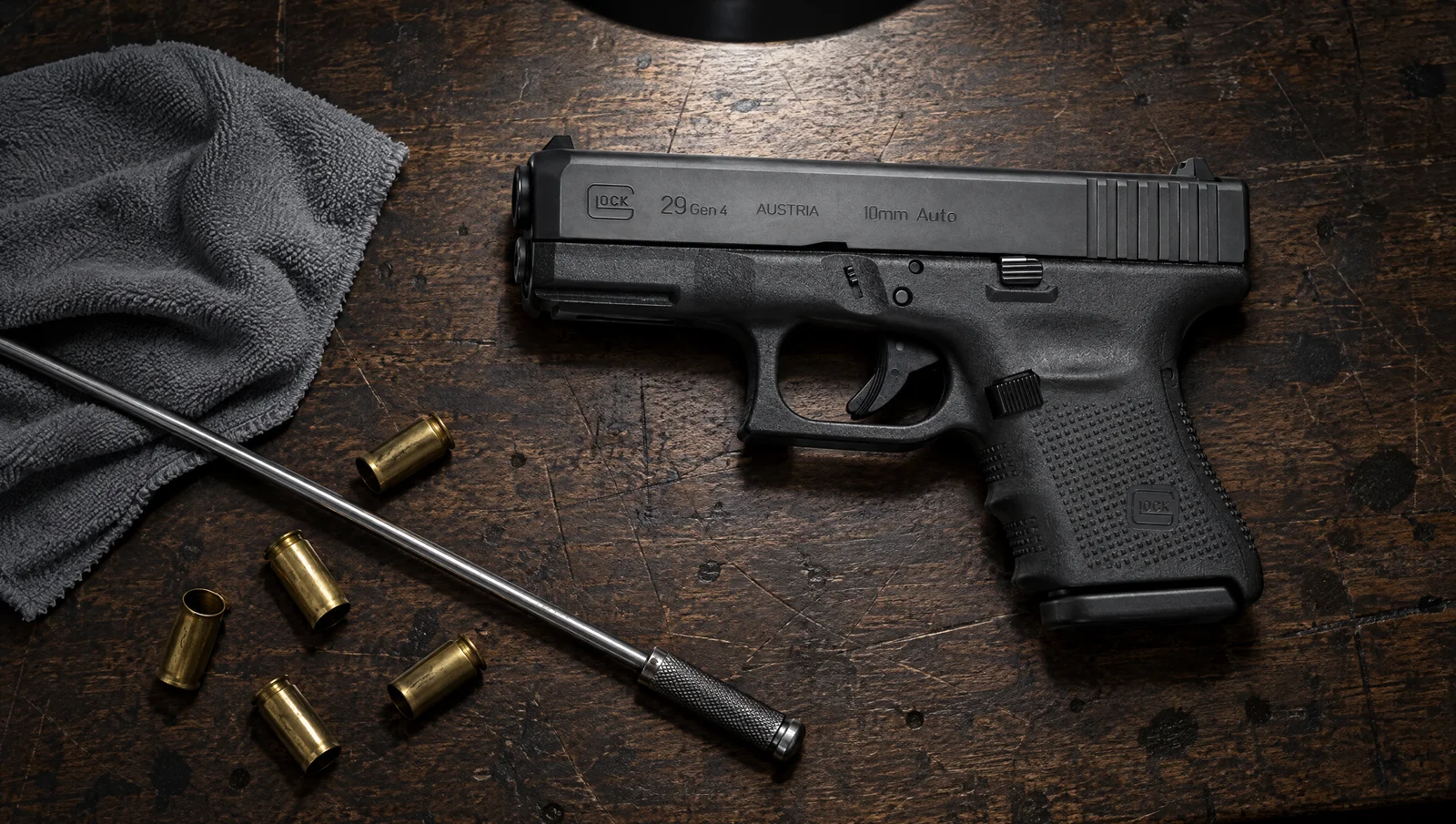 Glock 29 Gen4 10mm pistol lying on a dark oak workbench under a work lamp next to a cleaning rod, a gray cloth and empty 10mm brass