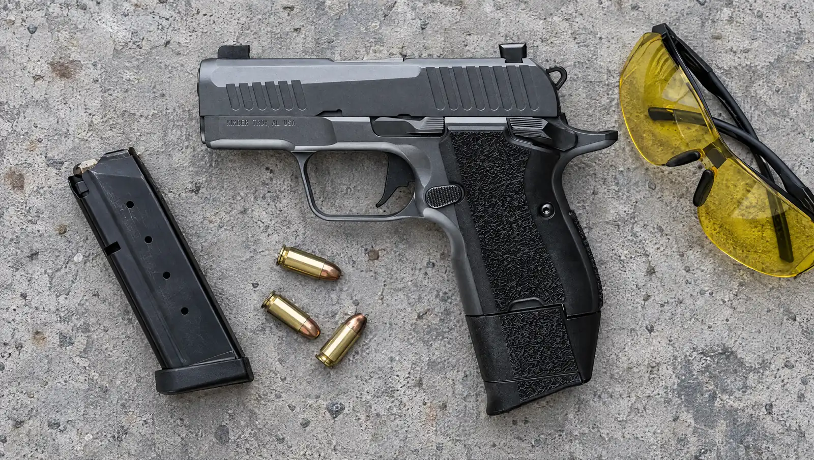 Kimber CDS9 Classic 9mm in gray KimPro II with black stippled grips lying on a concrete range bench beside a spare magazine, 9mm cartridges and yellow shooting glasses