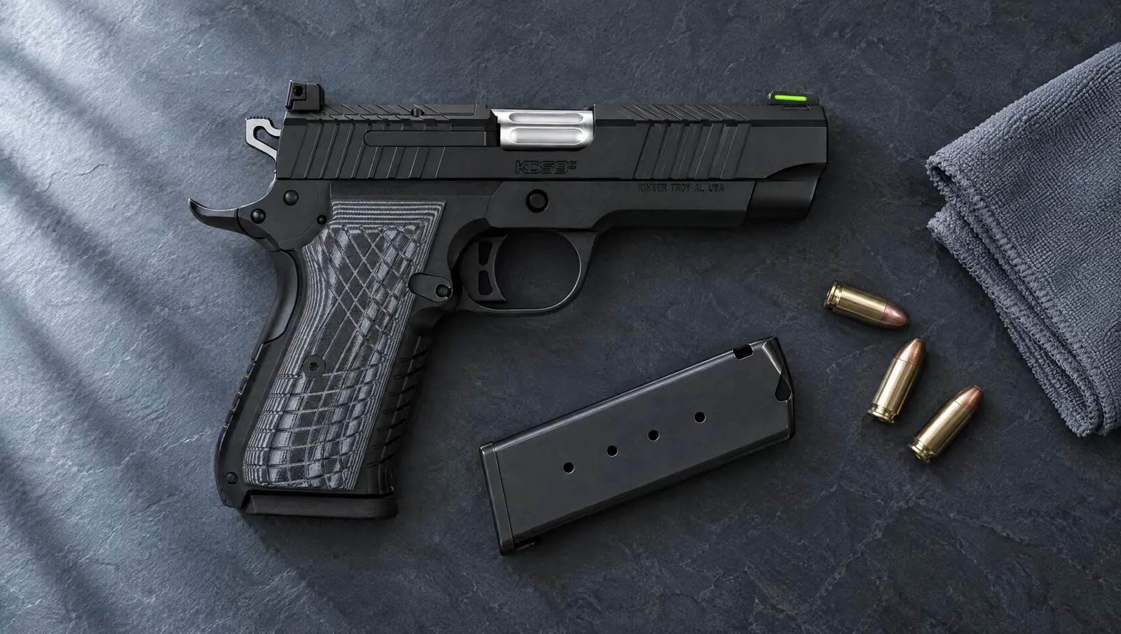 Kimber KDS9c 9mm double stack 1911 in black with a KDS9c slide logo, KIMBER TROY AL USA frame marking and crosshatch G10 grips lying on a slate countertop beside a spare magazine, 9mm cartridges and a microfiber cloth