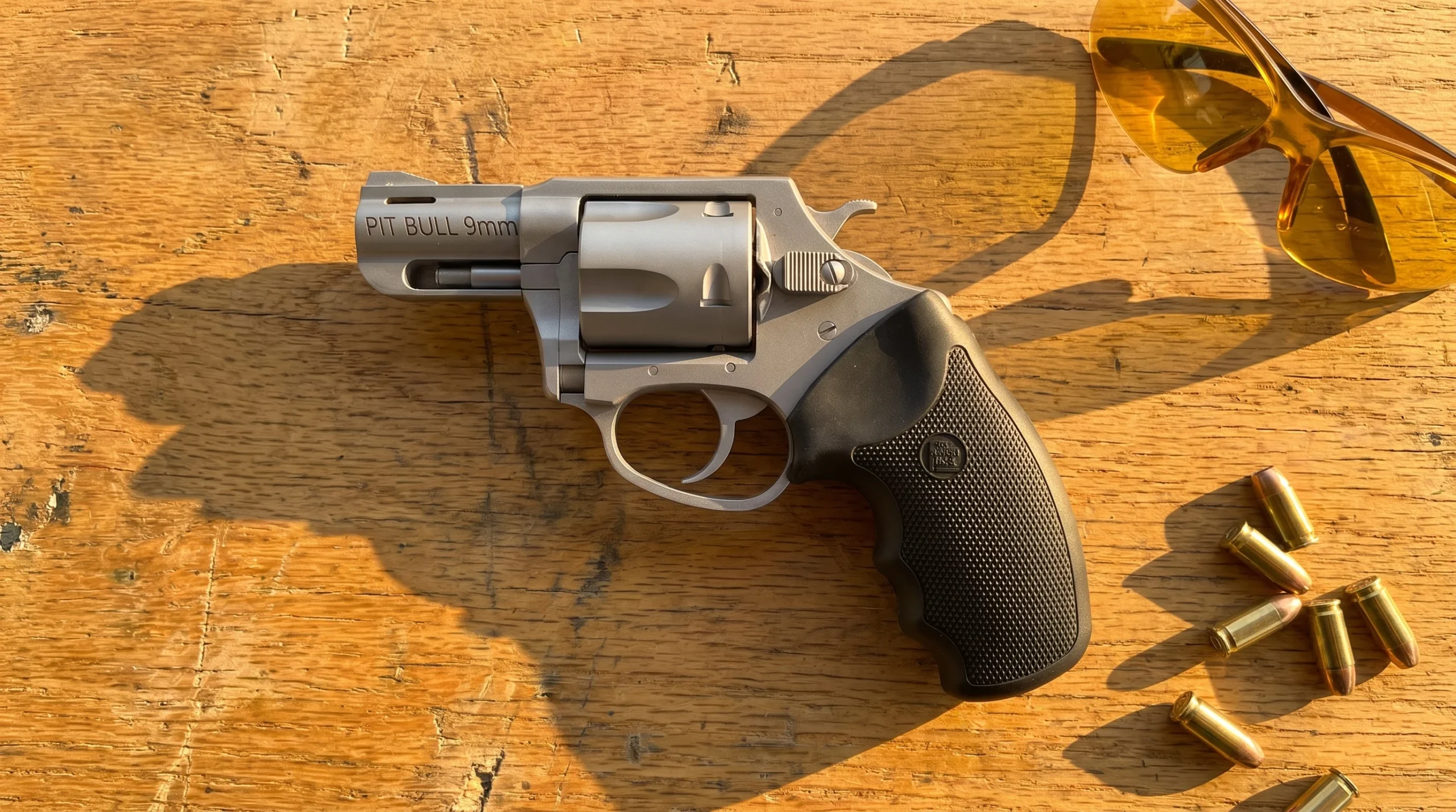 Charter Arms Pitbull 9mm stainless revolver on an oak shooting bench with 9mm cartridges and amber shooting glasses