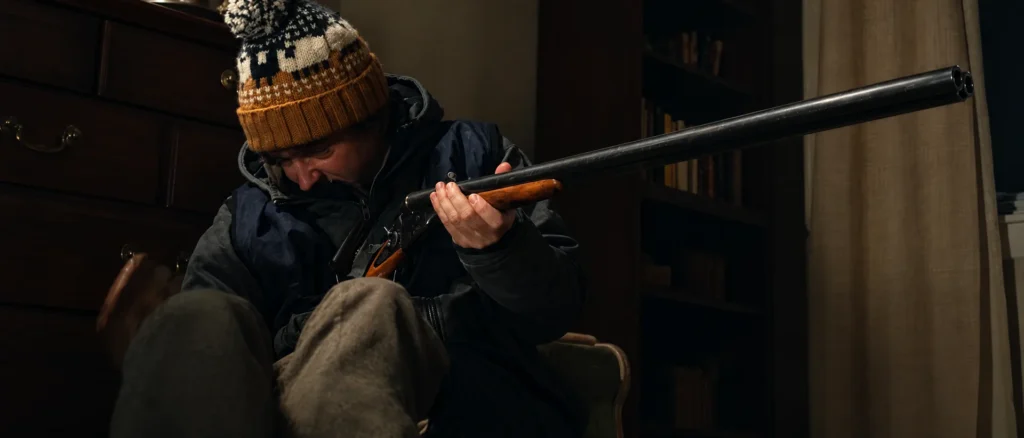 Bryan in a knit hat holding the hammer double-barrel shotgun in a dark study in Resident Evil 2026