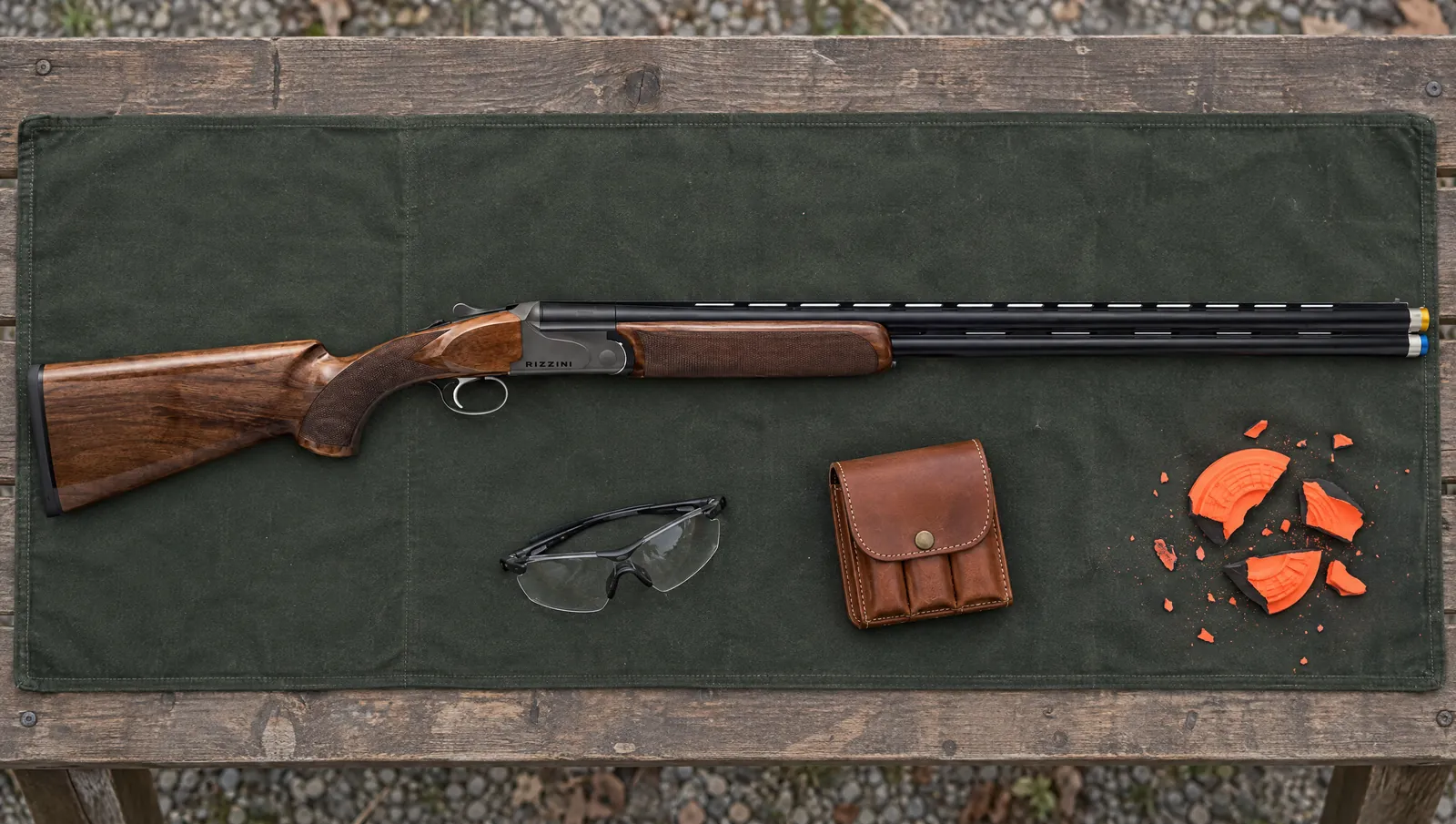 Rizzini BR110 Sporting 12 gauge with a smoke gray Cerakote receiver marked RIZZINI, 32 inch barrels with ventilated side ribs and extended chokes, lying on a green canvas mat on a clays shooting station beside shooting glasses, a choke pouch and broken orange clays