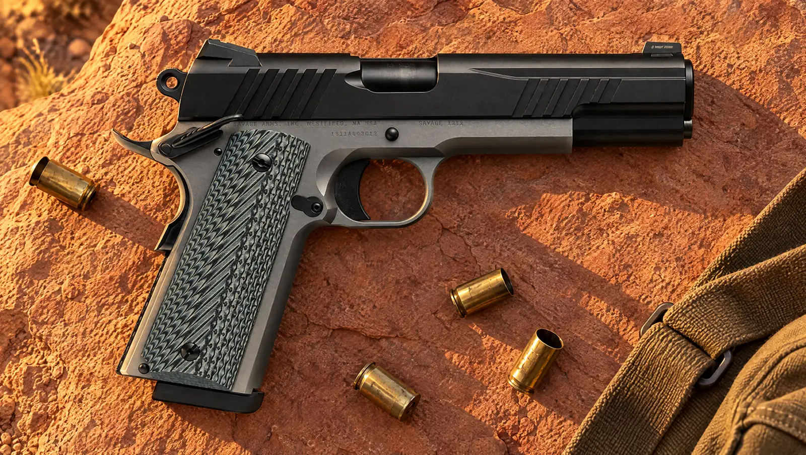 Savage 1911 two-tone .45 ACP with a black nitride slide, stainless frame and gray G10 grips lying on red sandstone at sunrise beside spent .45 brass