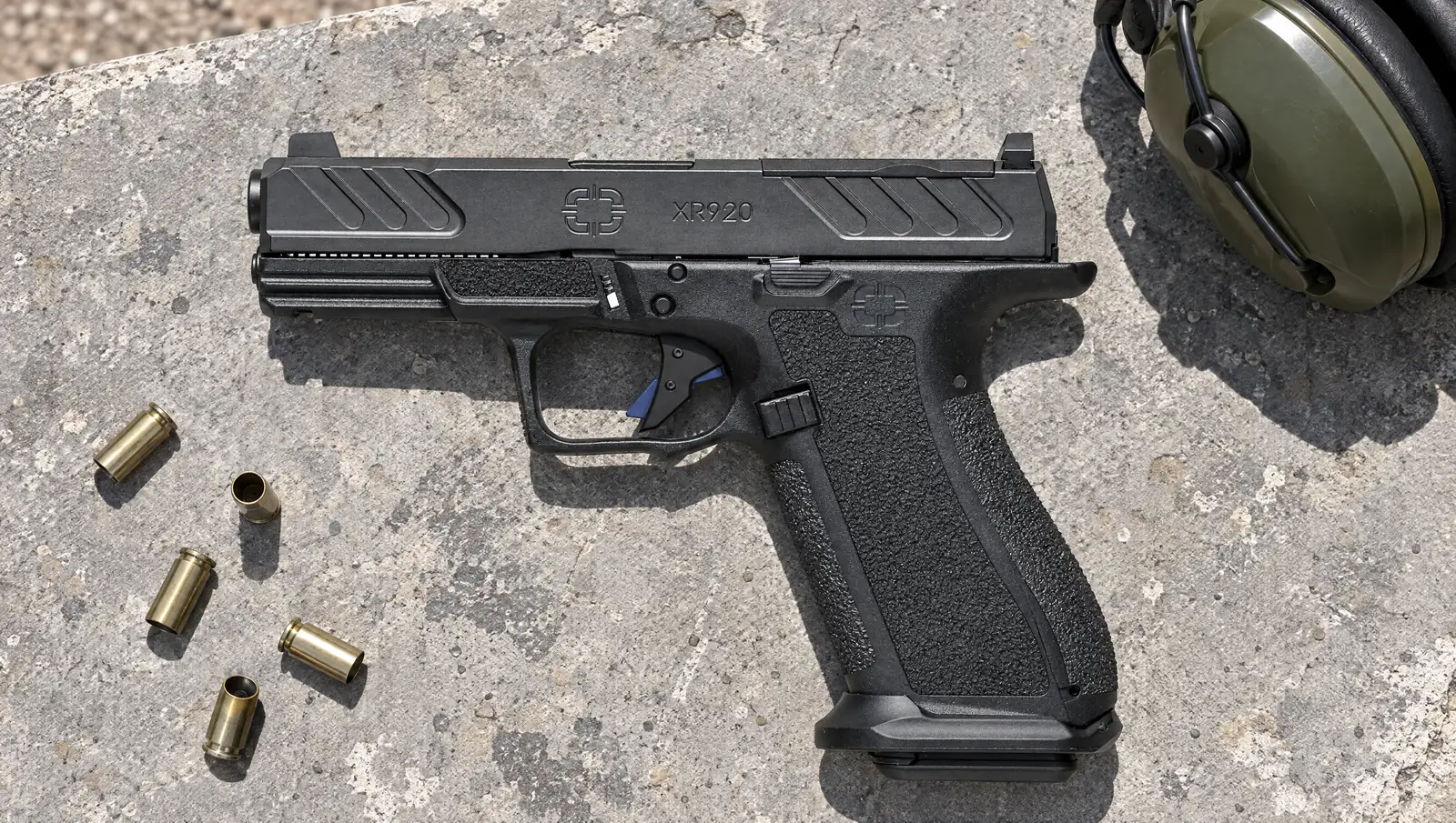 Shadow Systems XR920 LE 9mm pistol with a blue trigger safety lying on a concrete range bench next to ear protection and spent 9mm brass
