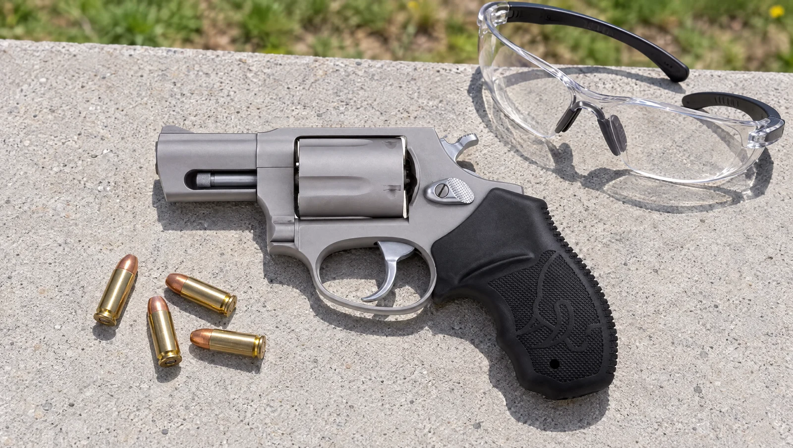 Matte stainless Taurus 905 9mm revolver lying on a concrete range bench next to loose 9mm cartridges and shooting glasses