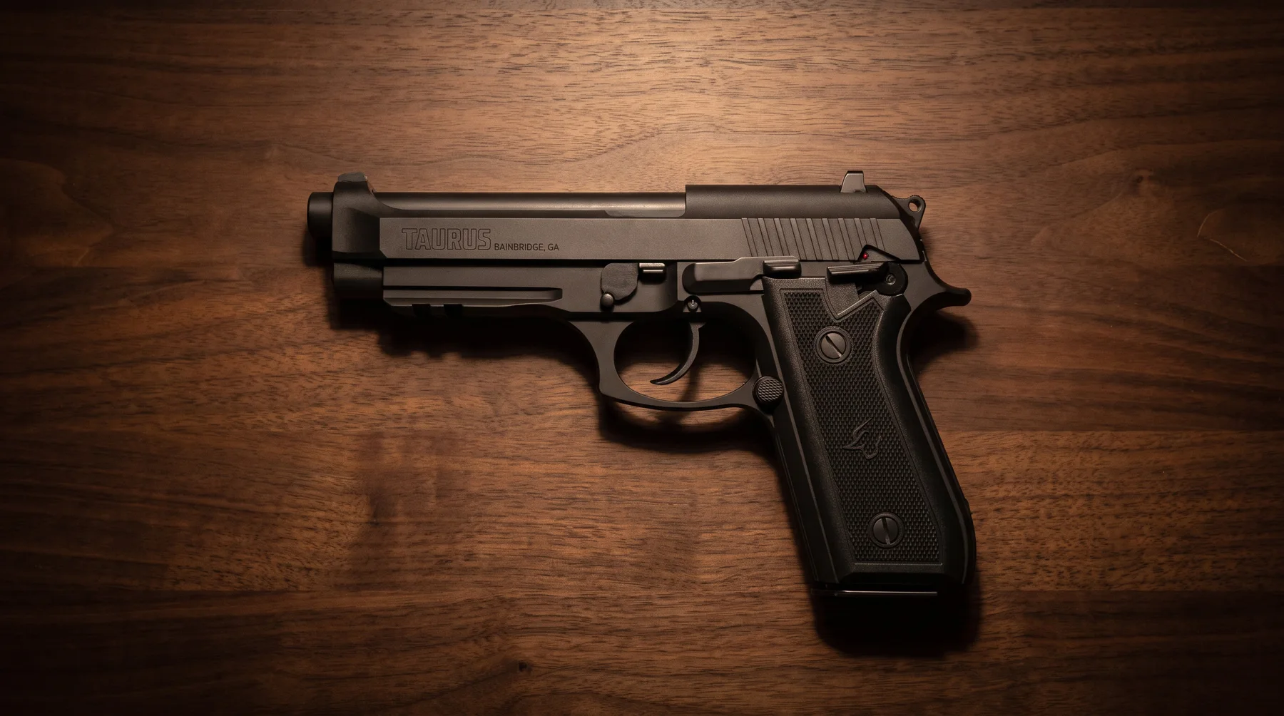 Taurus 92 matte black 9mm pistol lying on a walnut desk under warm lamp light, seen from above