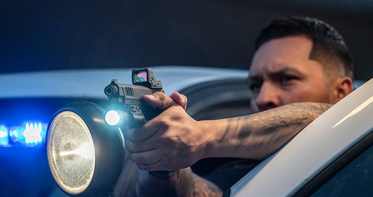 Shooter aiming a pistol with a Trijicon RMR HD and a weapon light beside a car with blue emergency lights