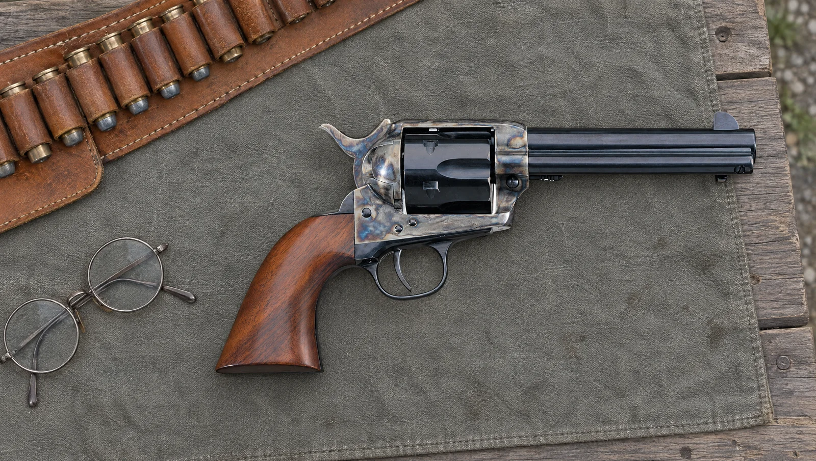 Uberti 1873 Cattleman .45 Colt revolver with a 4.75 inch barrel, case hardened frame and walnut grip lying on a gray canvas cloth beside a leather cartridge belt and round steel eyeglasses