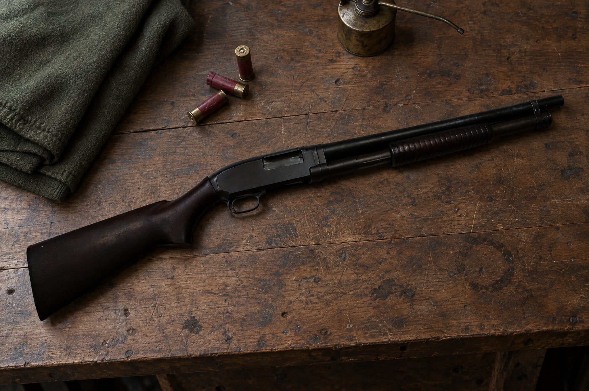 Winchester Model 12 riot shotgun lying on a wooden armory workbench with red shotshells, a wool blanket and an oil can