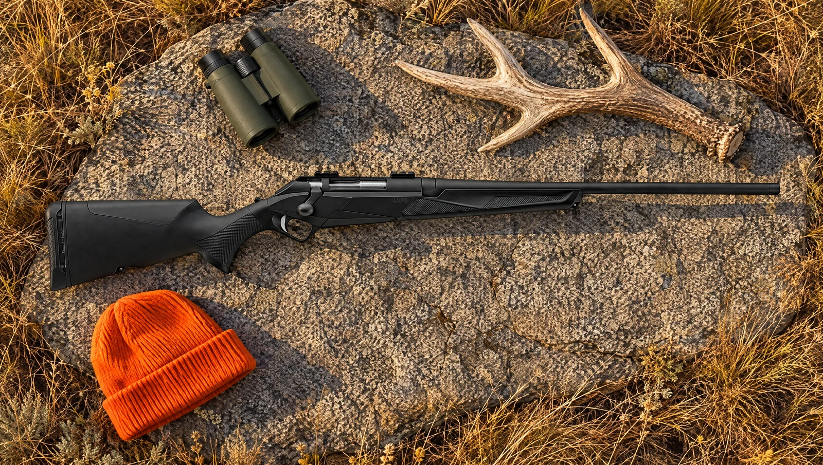 Benelli Lupo bolt-action rifle lying flat on a granite slab in autumn prairie grass beside binoculars, an elk antler shed and a blaze orange hat
