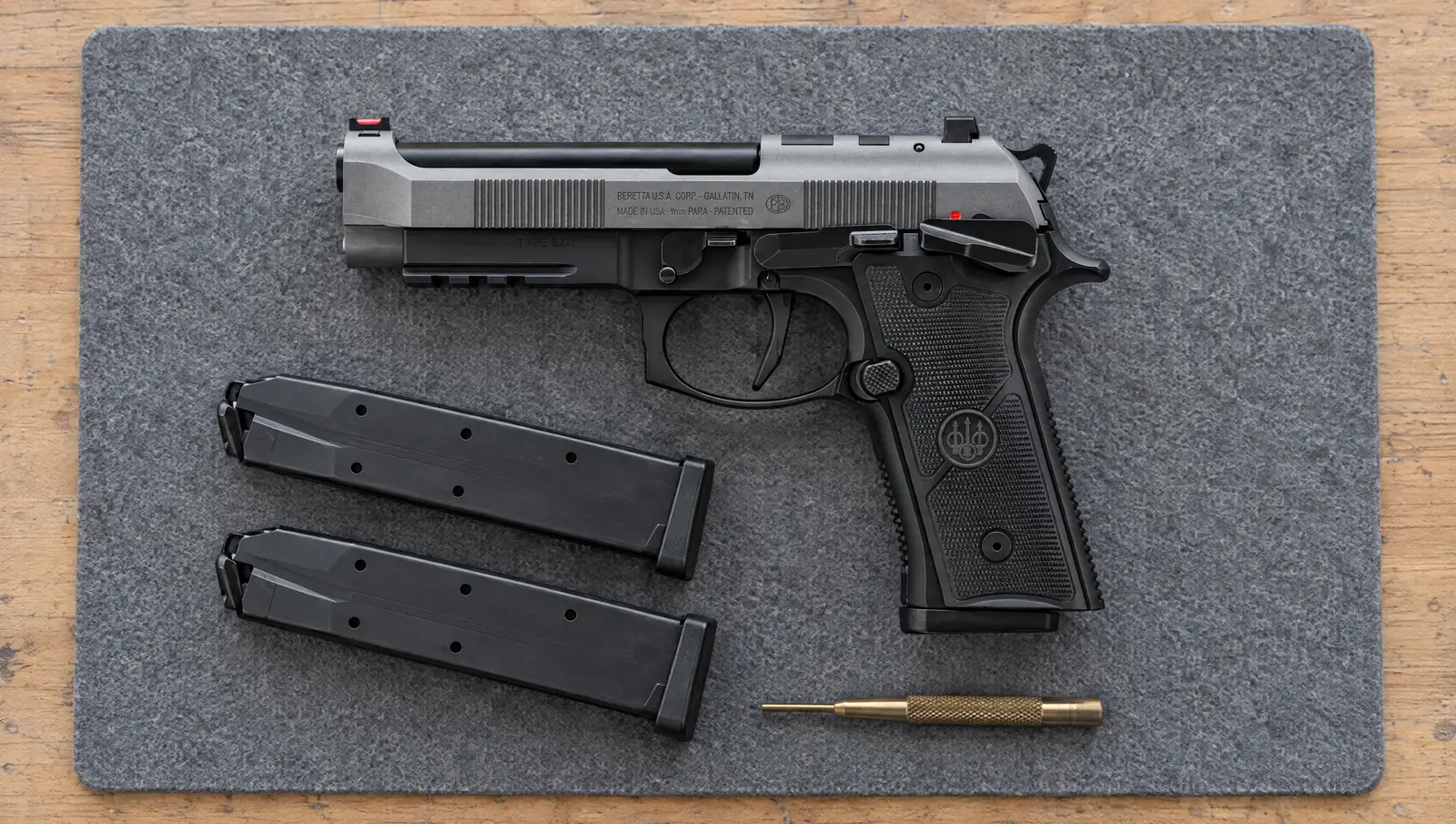 Beretta 92XI SAO 9mm pistol with a gray slide marked Beretta U.S.A. Corp. Gallatin, TN and a black frame with a frame-mounted safety, lying on a bench mat beside two magazines and a brass punch