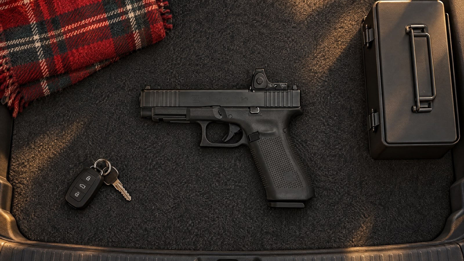 Glock 47 MOS pistol lying flat on the carpet of an SUV cargo area beside a closed steel lockbox, car keys and a plaid blanket