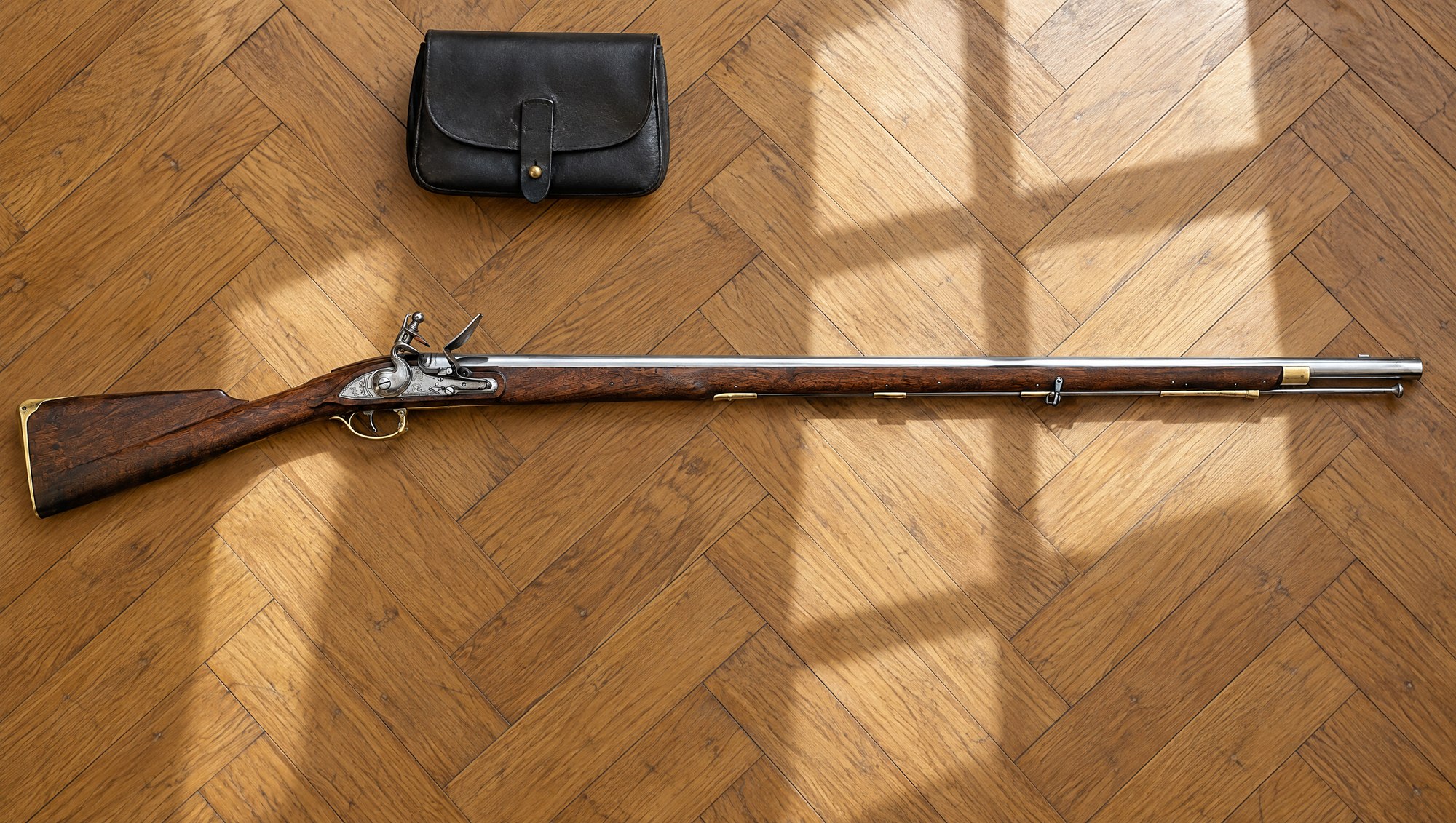 Brown Bess musket: a Pedersoli Brown Bess flintlock lying flat on a herringbone oak floor beside a black leather cartridge pouch