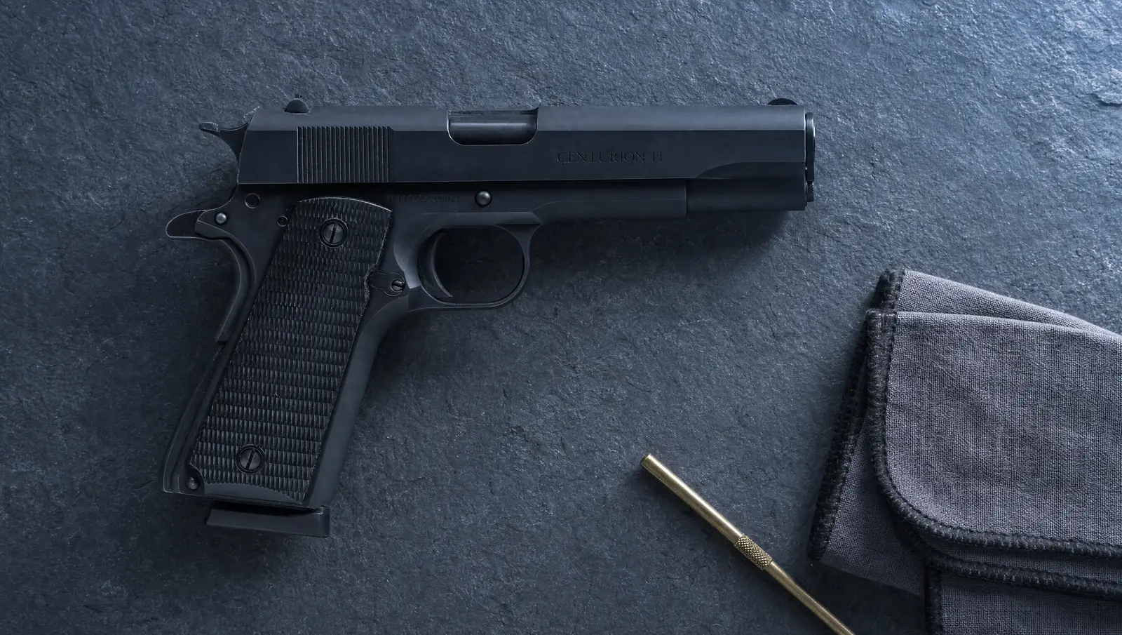 Black Century Arms Centurion 11 .45 ACP 1911 with checkered black grips and Centurion 11 slide marking lying on a dark slate tabletop beside a brass cleaning rod and a gun cloth
