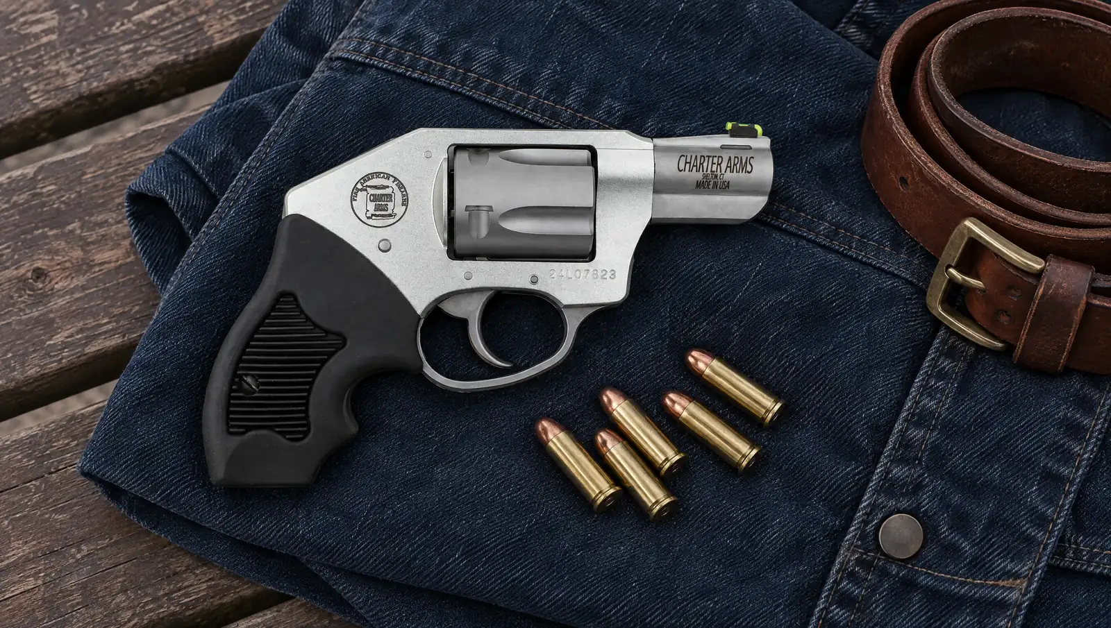 Charter Arms Off Duty 53811 .38 Special revolver with a clear anodized aluminum frame, enclosed hammer, green LitePipe front sight and black rubber grip lying on a folded denim jacket beside a leather belt and five .38 Special cartridges