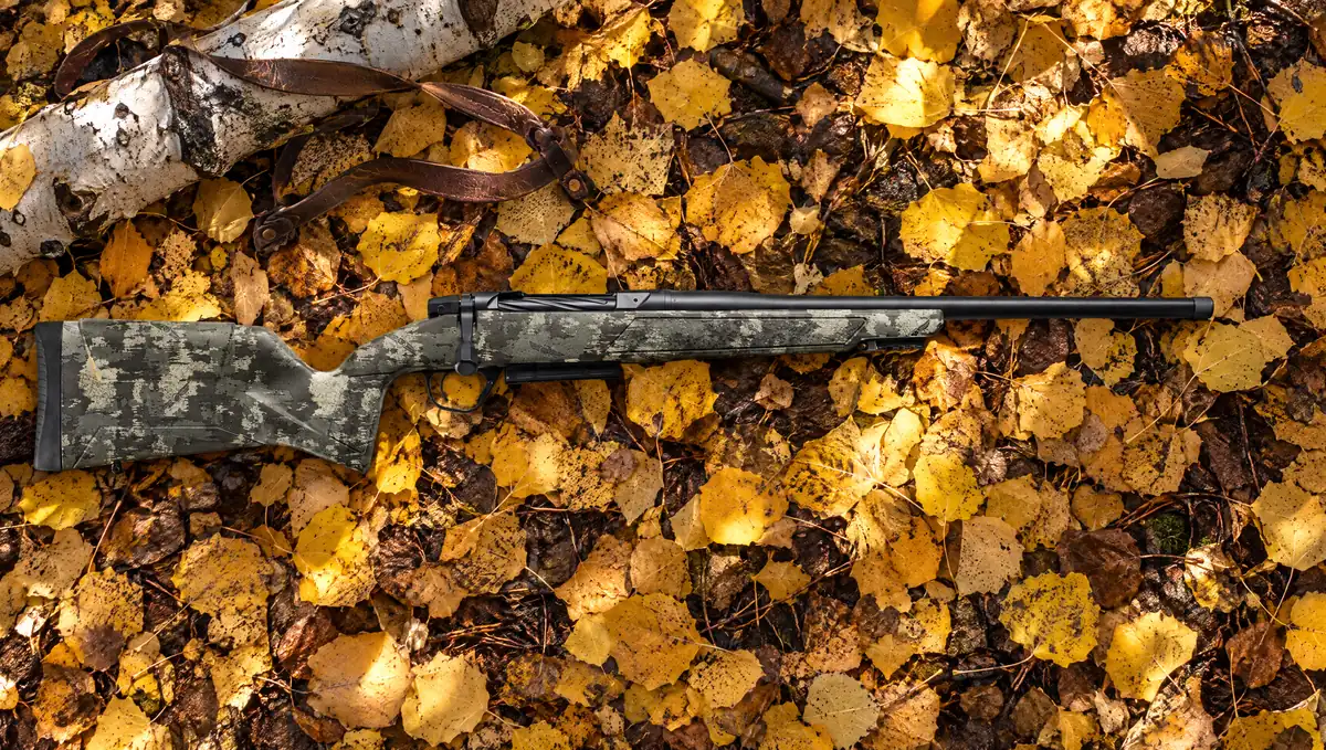 Christensen Arms Evoke Hunter bolt-action rifle with Brush Camo stock and Black Cerakote barrel lying on fallen golden aspen leaves beside an aspen log