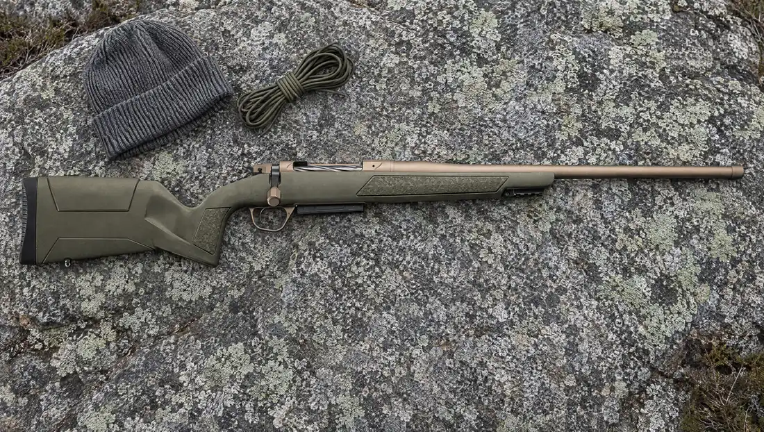 Christensen Arms Evoke bolt-action rifle with OD Green polymer stock and Smoked Bronze Cerakote barrel lying on lichen-covered granite beside a gray wool beanie and a coil of paracord