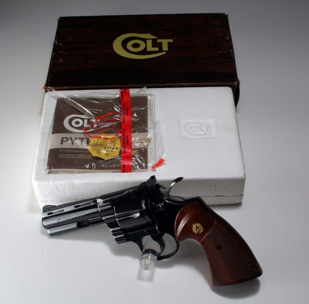 A blued four inch Colt Python in front of its woodgrain Colt box, foam insert and sealed instruction manual