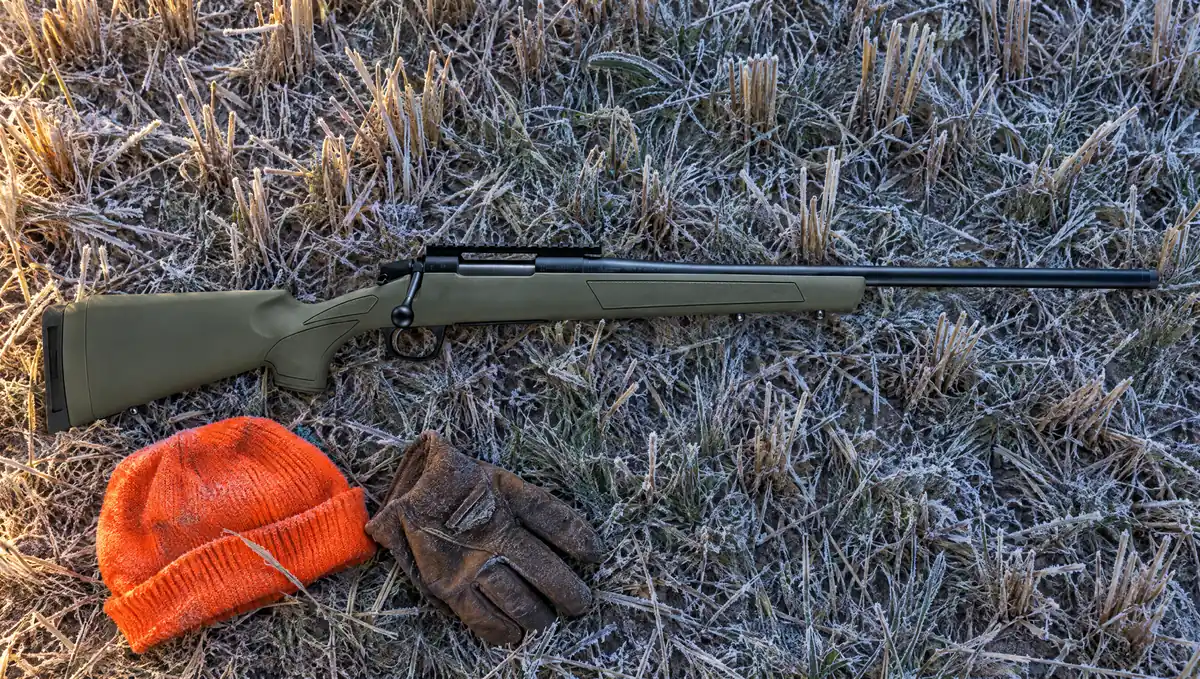 CVA Cascade bolt-action rifle with OD Green synthetic stock and Graphite Black Cerakote barrel lying on frosted wheat stubble beside a blaze orange knit cap and a leather glove