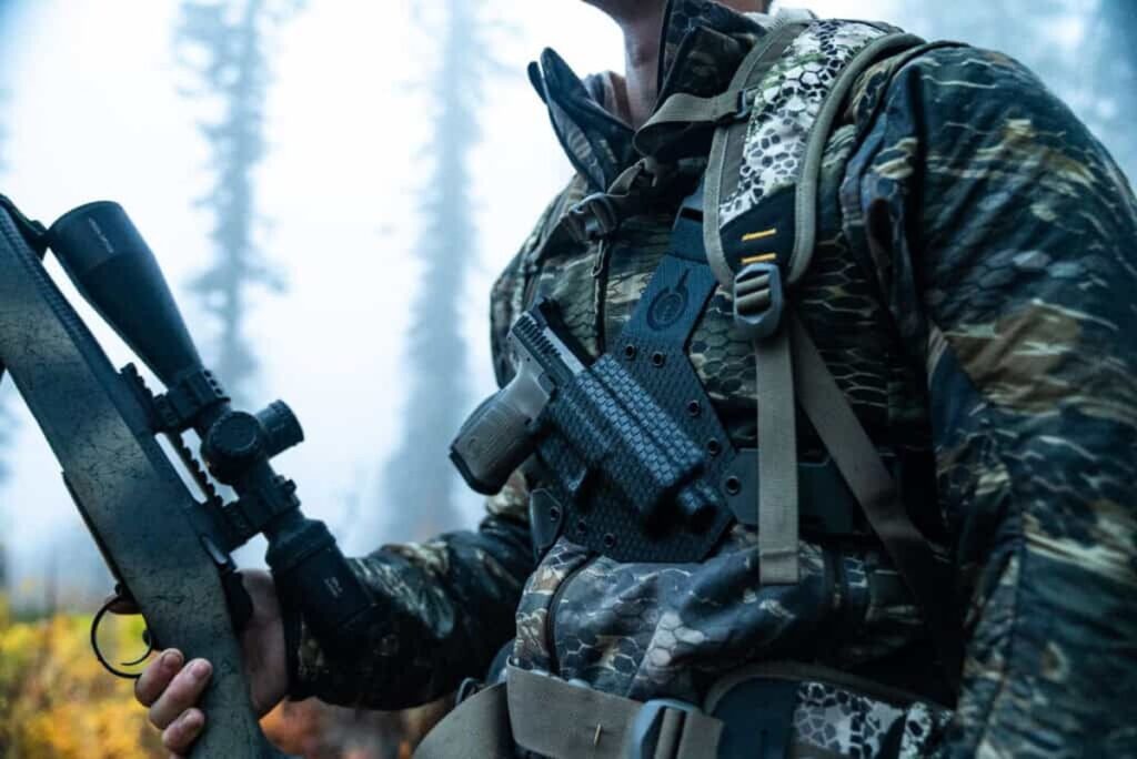 Hunter wearing a GunfightersINC Kenai chest holster with a scoped rifle in a misty forest