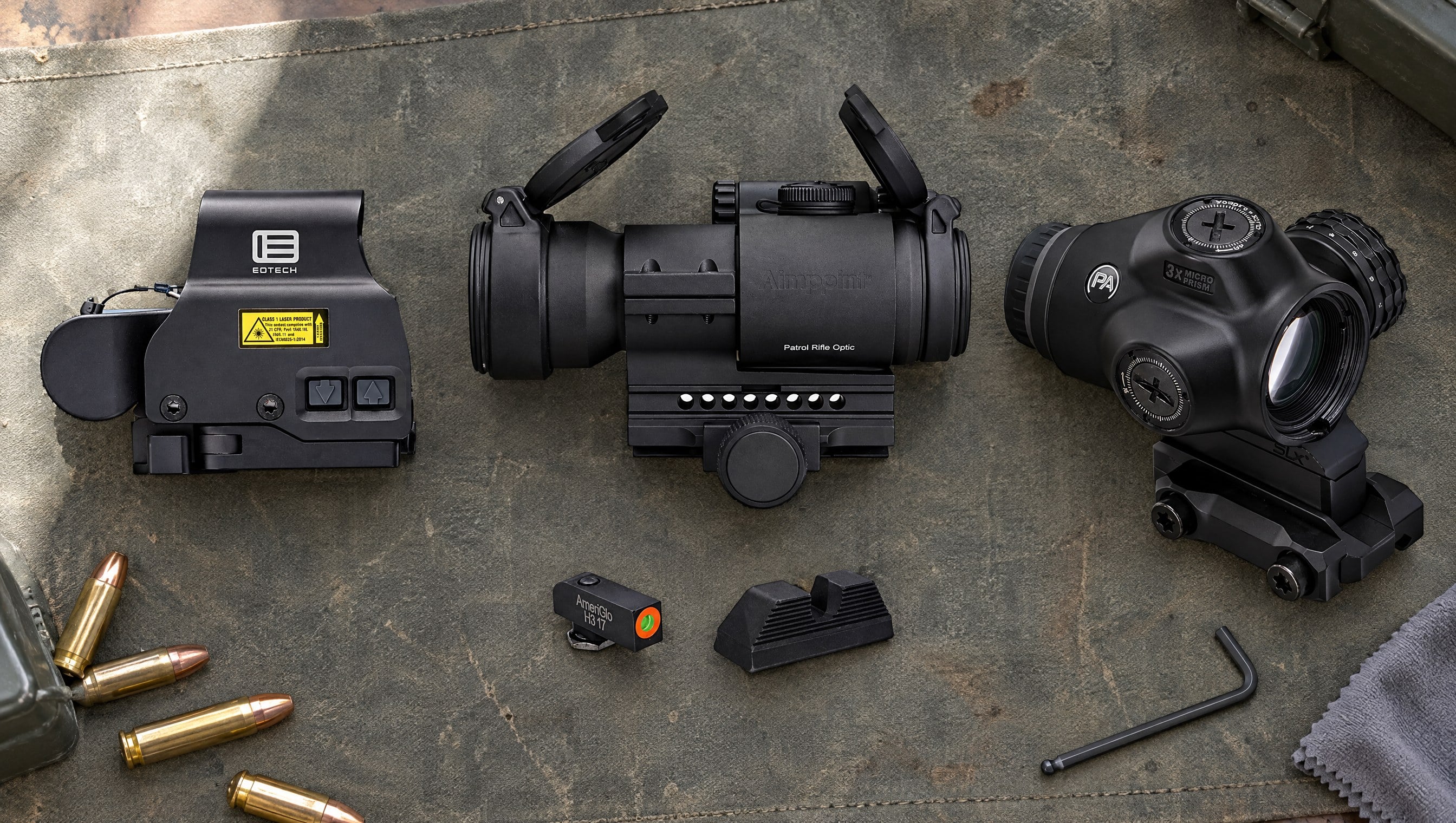 An EOTech holographic sight, an Aimpoint Patrol Rifle Optic red dot, a Primary Arms SLx 3x MicroPrism scope and an AmeriGlo front and rear night sight set laid out on a gray-green canvas shooting mat