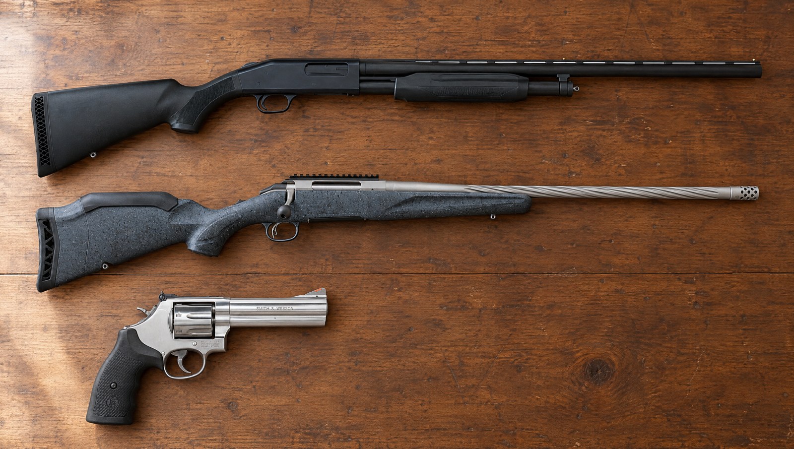 A Smith and Wesson 686 revolver, a Ruger American bolt-action rifle and a Mossberg 500 pump shotgun on a walnut table