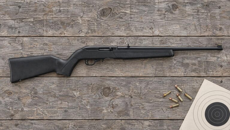 Ruger 10/22 Carbine model 32002 with black polymer stock lying on a wooden shooting bench beside .22 LR cartridges and a paper target