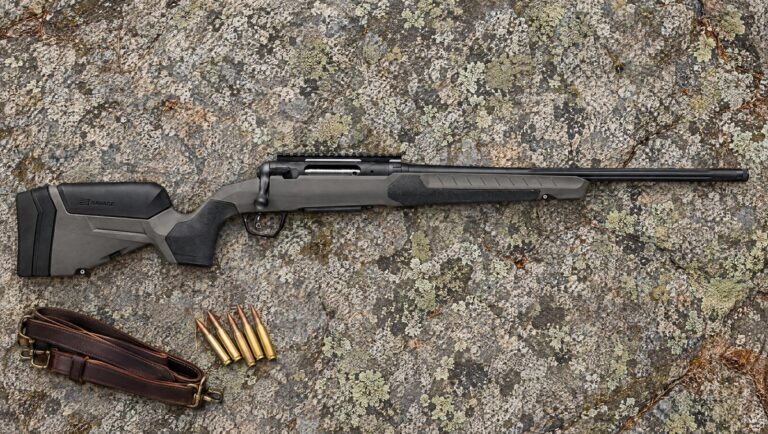 Savage 110 Core Hunter bolt-action rifle with gray synthetic stock and bare scope rail lying flat on lichen-covered granite beside rifle cartridges and a leather sling