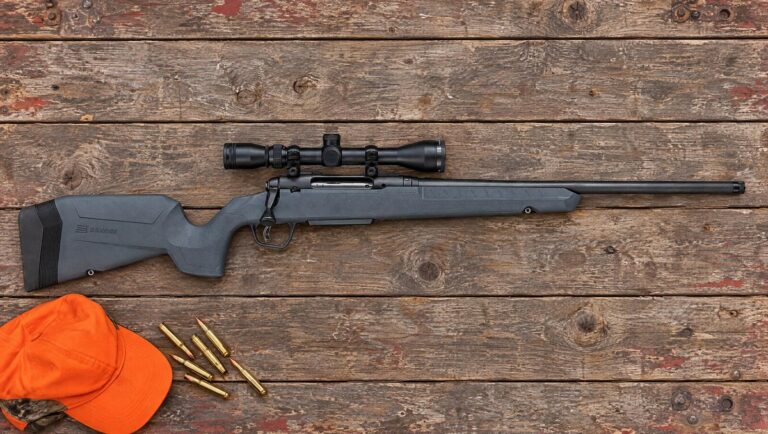 Savage Axis 2 XP bolt-action rifle with gray synthetic stock and factory 3-9x40 scope lying flat on a barn-wood table beside rifle cartridges and an orange hunting cap