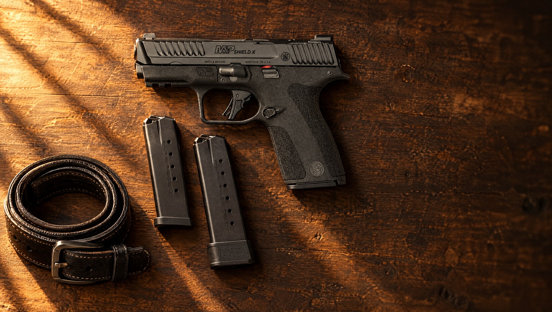 Smith & Wesson M&P Shield X 9mm lying on a walnut table beside its 13 and 15 round magazines and a leather belt