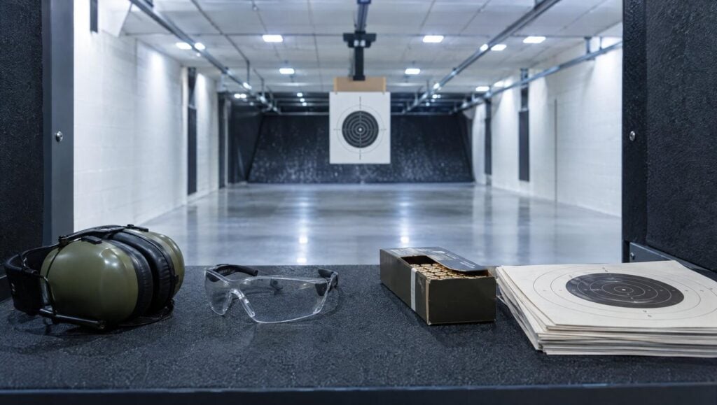 Empty indoor shooting range lane with a paper target downrange and ear muffs, glasses and ammo on the bench