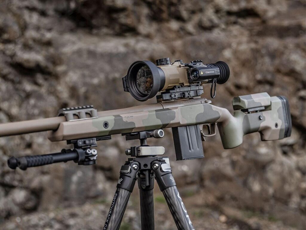 Tan iRay Rico Hybrid 640 75 mm thermal sight with an ILR rangefinder module mounted on a camo bolt-action rifle on a tripod
