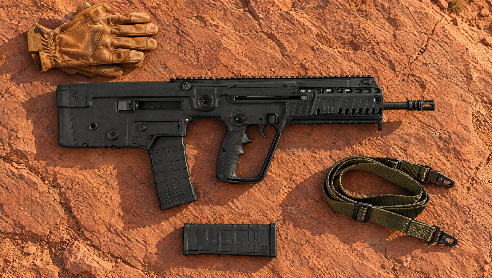IWI Tavor X95 5.56 bullpup rifle lying flat on red sandstone beside leather gloves, a spare magazine and an olive sling
